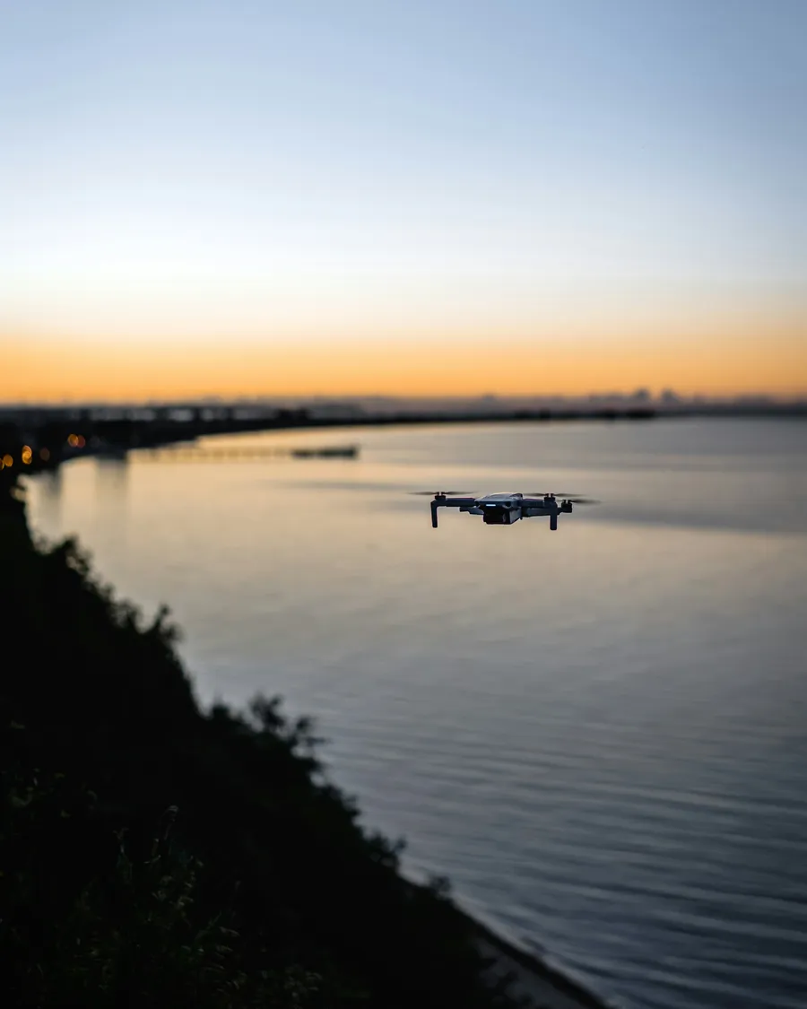 A drone hovering low over still water at dusk, with a city skyline faint on the horizon